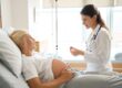 Pregnant woman lying in bed and a doctor checking her abdomen in a hospital room, both smiling.