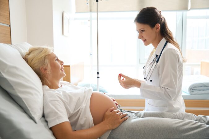 Pregnant woman lying in bed and a doctor checking her abdomen in a hospital room, both smiling.