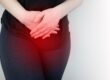Person standing against a light wall, hands clasped over the abdomen, suggesting stomach pain or discomfort.
