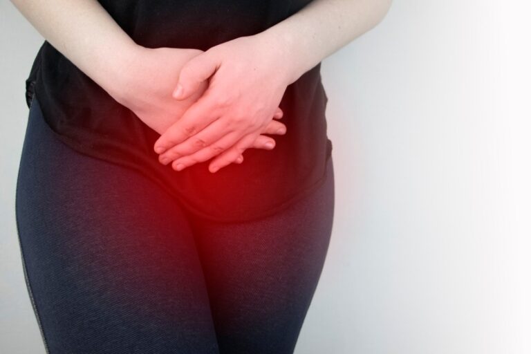 Person standing against a light wall, hands clasped over the abdomen, suggesting stomach pain or discomfort.