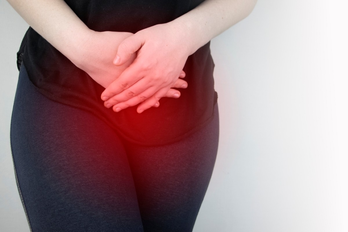 Person standing against a light wall, hands clasped over the abdomen, suggesting stomach pain or discomfort.