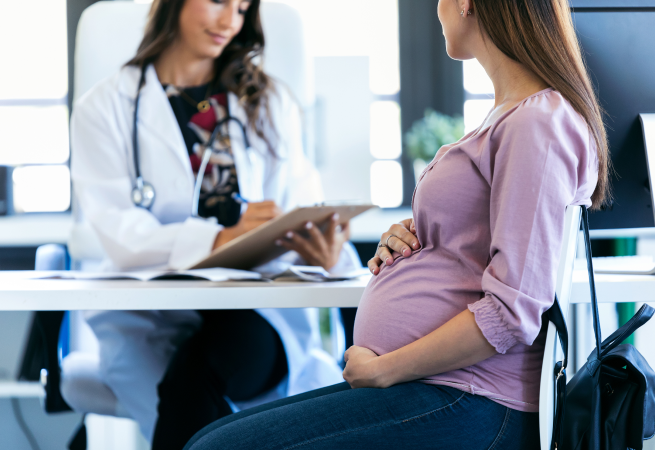 henderson-recruitment-image5 Pregnant woman sitting at a clinic table, holding her belly while a doctor reviews notes on a clipboard.