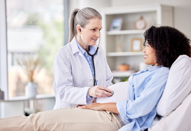 Doctor uses a stethoscope to listen to a patient’s chest during a check-up in a bright clinic room, both smiling at each other.