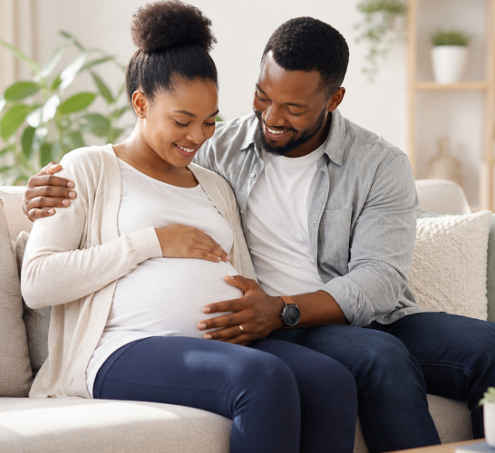 Pregnant woman sits on a sofa with her partner, both smiling as they rest a hand on her baby bump in a cozy living room setting.