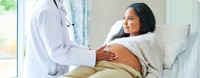 hera-benefits-img (1) Pregnant woman in a hospital bed as a doctor palpates her baby bump during a prenatal exam.
