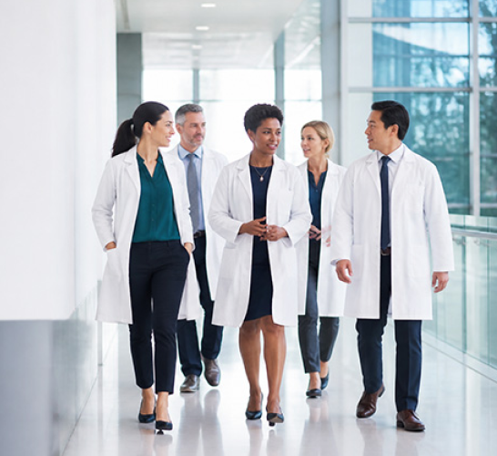 A diverse group of five doctors in white coats walking down a bright hospital corridor, engaged in conversation.