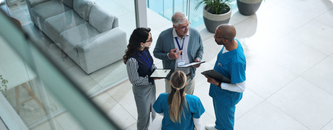 reason-to-join-img (3) Healthcare professionals in scrubs and colleagues stand in a bright lobby discussing a tablet and documents.
