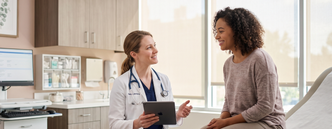 Doctor in a white coat with a stethoscope shows a tablet to a smiling patient in a bright clinic exam room.